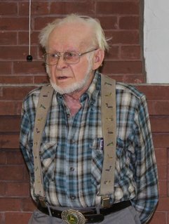 Elderly man with white hair, glasses, and plaid shirt stands against a brick wall.